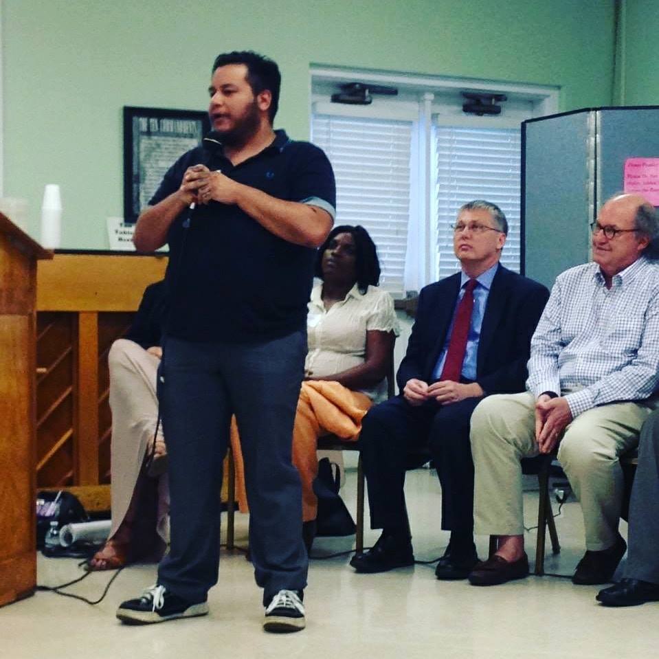 Jack Knoxville is a brown skinned trans man seen wearing a black polo style shirt with jeans and sneakers while holding a microphone during a campaign event in Knoxville, TN