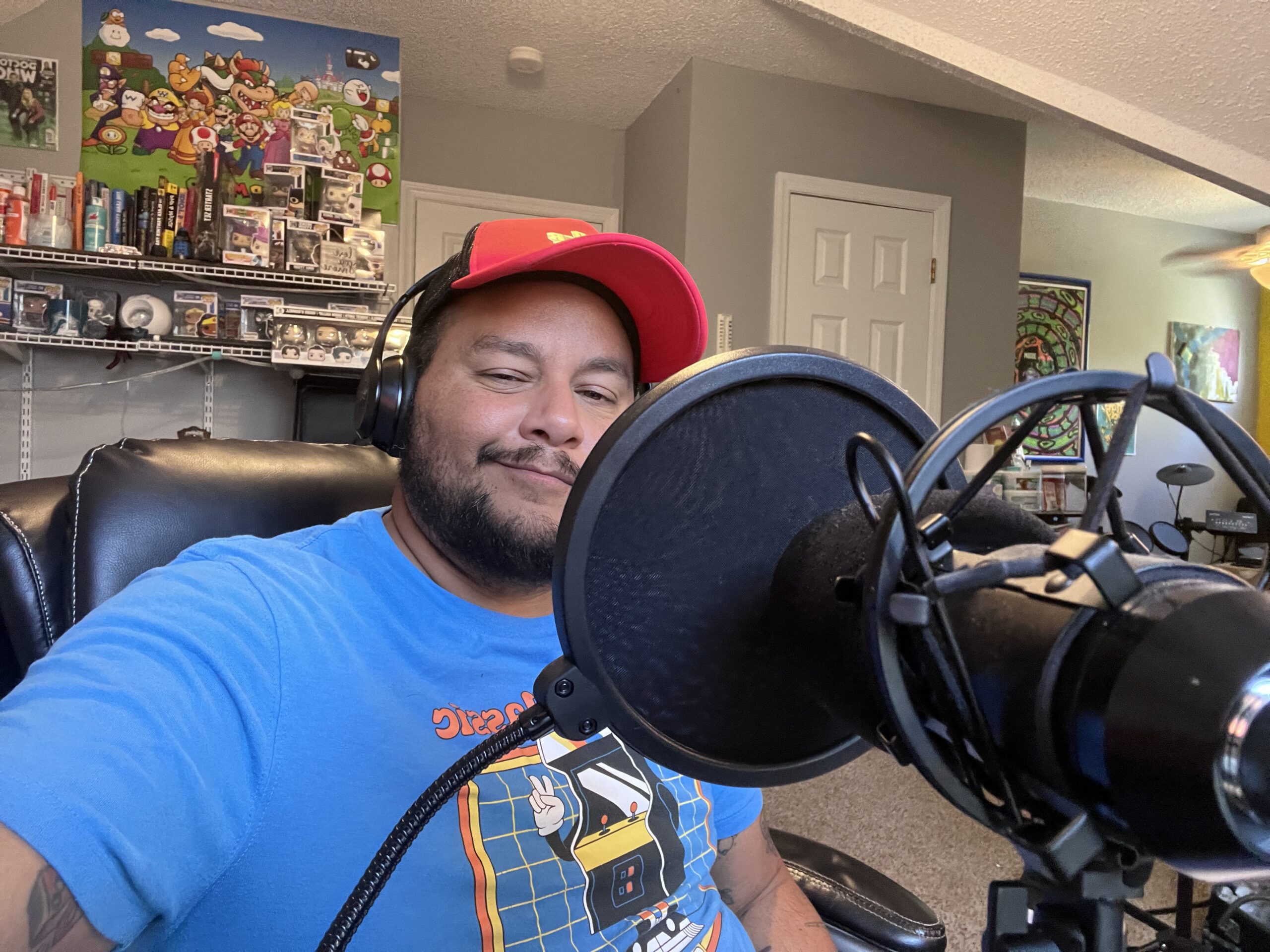 Jack is a medium brown skinned afro latino shown wearing a blue t-shirt and red hat, and smiling behind his microphone.